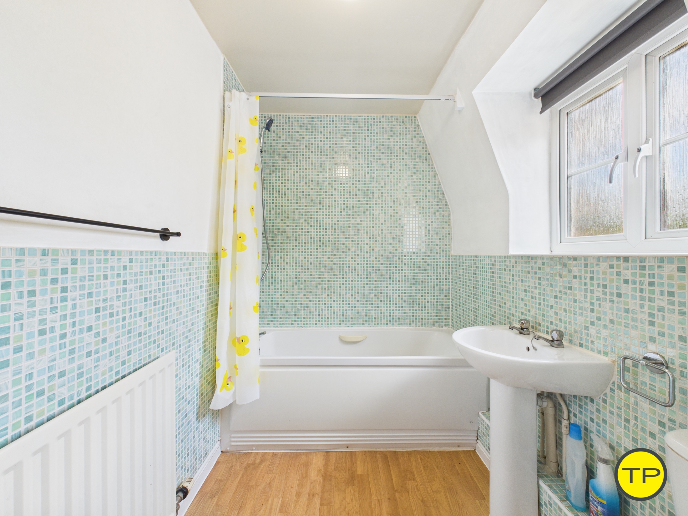 The bathroom features bright white walls and a light oak-effect vinyl floor. Stylish green mosaic splash backs add a pop of colour around the wet areas. The suite includes a white WC, hand basin, and a bath with a thermostatic shower above. There is also an obscure glazed window overlooking the rear of the home.
